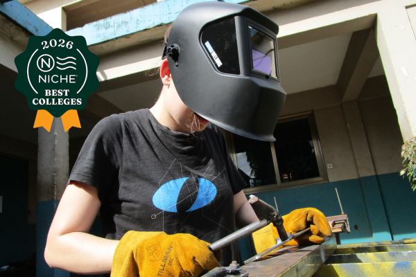 An Olin College of Engineering student is wearing a welder's mask and black t-shirt with the Olin "O" logo in blue across the middle.